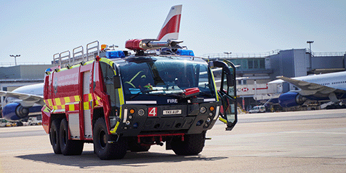 A photo of an airport specialised fire engine driving on the airfield, with planes in the background