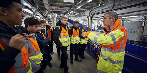A photo of a LGW colleague talking to a group of teenagers in high vis jackets in the backrooms of Gatwick Airport