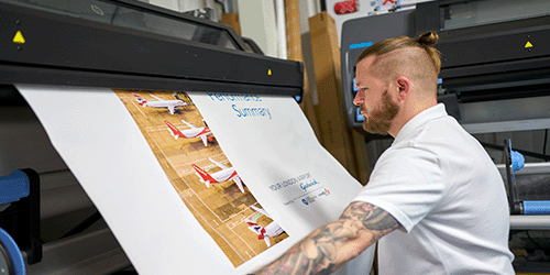 Photo of the print room of an industrial scale printing and sign making business, with a man holding a London Gatwick poster and Gatwick airport signs in the background