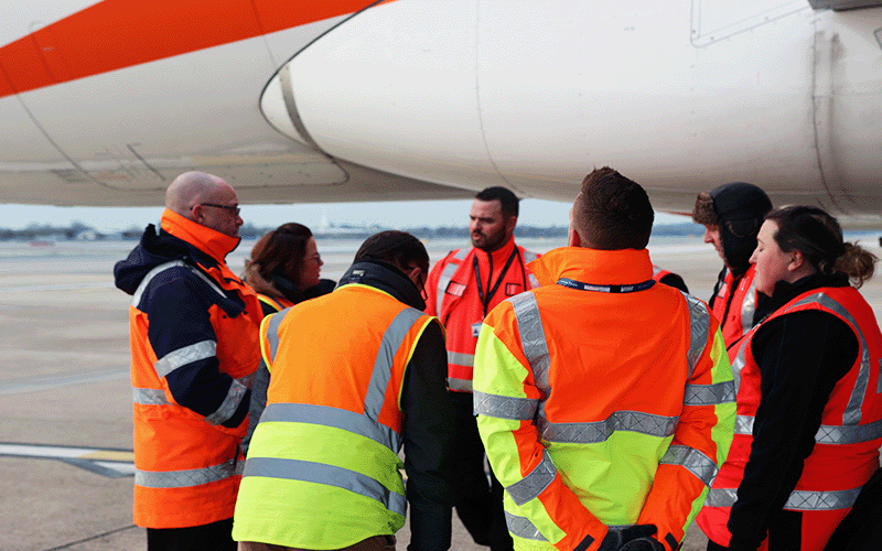 A photograph of a group of 7 Gatwick and easyJet colleagues in high-vis jackets gathered underneath the wing of an easyJet aircraft, having a discussion