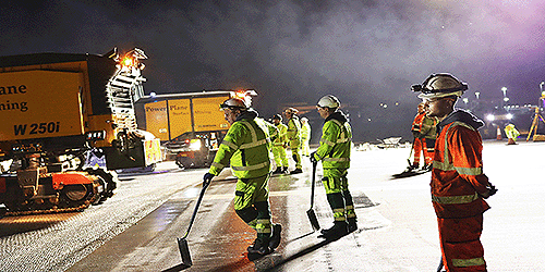 A photograph of a group of runway resurfacing engineers in high vis vests and hard hats, holding shovels, standing on London Gatwick's main runway at night. There is also large machinery.