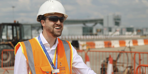 Profile photograph Sam Hayter in high vis vest with pier 6 bridge and a JCB digger behind him