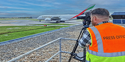 A photo of a man in a high vis vest with a camera and tripod filming an Emirates a380 taxiing past