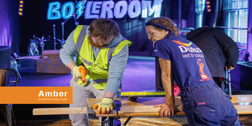 An image, via the Amber Foundation, of a man sawing a plank of wood, supervised by a tutor