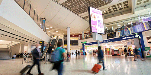 Long exposure photograph of passengers walking through London Gatwick's North Terminal, with a large advertising screen in the centre