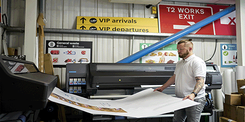 Photo of the print room of an industrial scale printing and sign making business, with a man holding a London Gatwick poster and Gatwick airport signs in the background