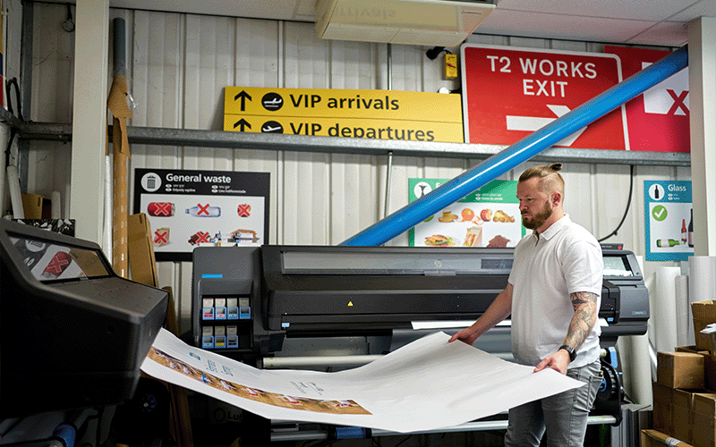 Photo of the print room of an industrial scale printing and sign making business, with a man holding a London Gatwick poster and Gatwick airport signs in the background